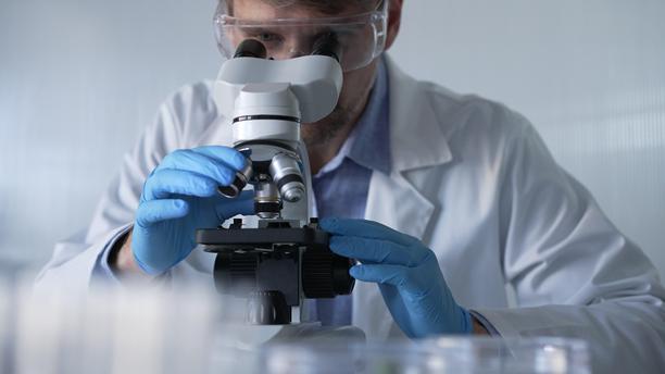 穿着蓝色手套和防护眼镜的男性科学家研究员正在现代实验室中使用显微镜，肖像视图。低调照明照片。科学与医学概念。图片下载