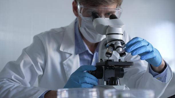 穿着蓝色手套、护目镜和医用口罩的男性科学家研究员正在现代实验室中使用显微镜，肖像视图。低调照明照片。科学与医学图片下载