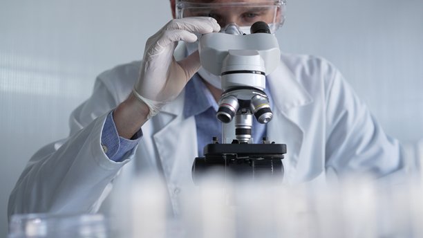 穿着白色手套、护目镜和医用口罩的男性科学家研究员正在现代实验室中使用显微镜，肖像视图。低调照明照片。科学与医学图片下载