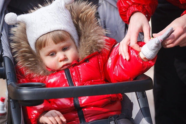 小女孩穿着红色冬季外套，坐在婴儿推车里，妈妈正在给她的手指戴上手套。横向图片。图片下载