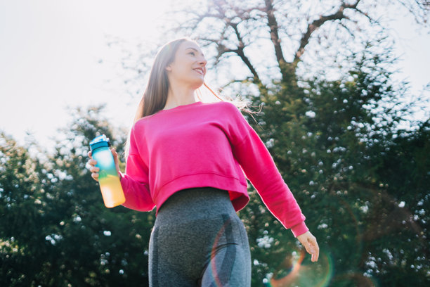 一位健康年轻的女性在公园锻炼后沐浴在阳光下喝水。她通过自己的例子强调了保持水分的重要性，同时这张照片散发出健康的生活方式和宁静感。图片下载