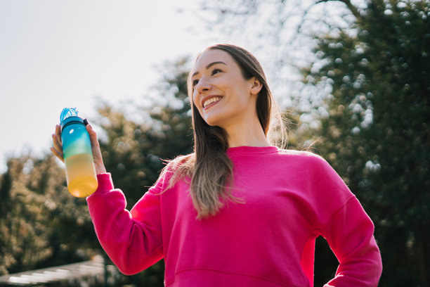 一位健康年轻女性在公园锻炼后沐浴在阳光下喝水。她通过自己的例子强调了保持水分的重要性，同时这张照片散发出健康的生活方式和宁静感。图片下载