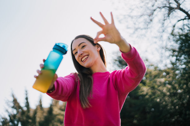一位健康年轻女性在公园锻炼后沐浴在阳光下喝水。她通过自己的例子强调了保持水分的重要性，同时这张照片散发出健康生活方式和宁静的氛围。图片下载