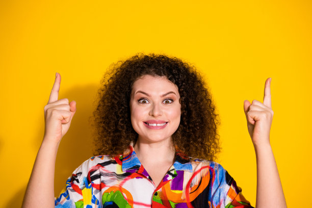 时尚的年轻女性在黄色背景下穿着色彩丰富的印花衣服，指向空白处的照片肖像。图片下载