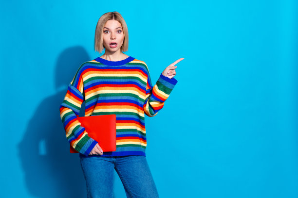 漂亮年轻女孩的照片肖像，手持笔记本电脑，指向空白处，表情惊恐，穿着时尚条纹服装，背景为蓝色。图片下载