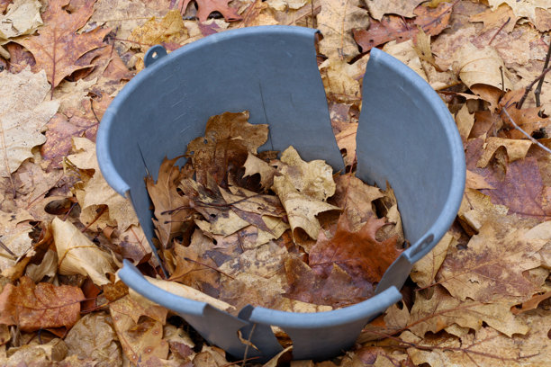 这张照片展示了一个受损的塑料桶，靠近地面，周围是落叶。桶是灰色的，表面有裂缝，显示出它的磨损。图片下载