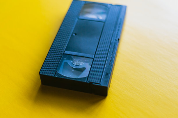 一张极简风格的VHS录像带在黄色背景上，形成了过时技术与现代美学之间的对比。照片强调了复古文化和怀旧设计的价值。图片下载