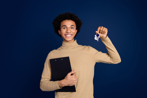 年轻男子的照片肖像，手持钥匙，穿着时尚的米色服装，背景为深蓝色。图片下载