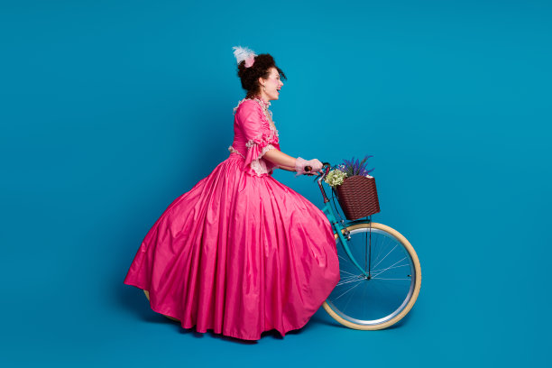 身穿华丽洛可可服装的美丽女士骑自行车的全身照片，背景为空旷的蓝色。图片下载