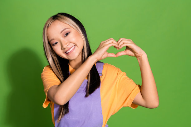 可爱的年轻女士在绿色背景下做心形手势，穿着时尚的紫色和黄色服装的照片肖像图片下载