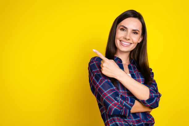 时尚的年轻女性在黄色背景下穿着格子衣服，指向空白处的照片肖像。图片下载