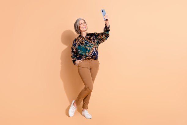 穿着印花上衣的快乐灰发人士在米色背景下用智能手机自拍的全身照片图片下载