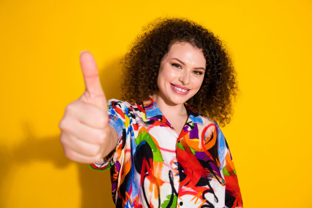 时尚年轻女士在黄色背景下竖起大拇指，穿着色彩鲜艳的印花服装的照片肖像图片下载