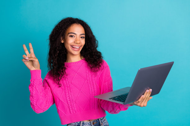 可爱的少女在青色背景下，穿着时尚的粉色服装，手持笔记本电脑，做出V字手势的照片肖像。图片下载