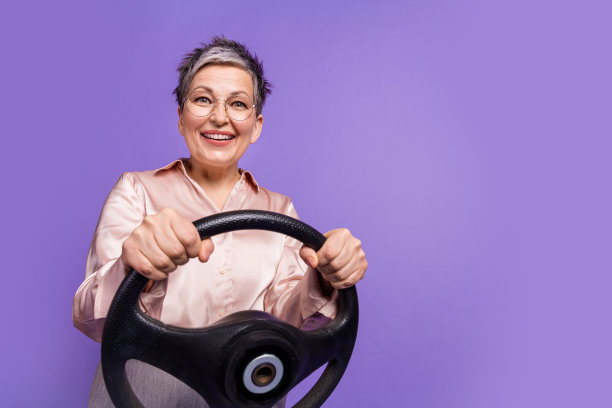 穿着时尚缎面服装的可爱退休女性手握方向盘的照片肖像，背景为紫色。图片下载