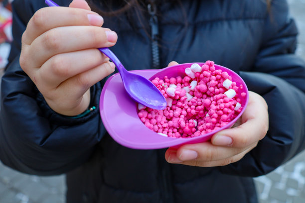 这张照片特写了一个孩子的手，手里拿着一个装有“迷你融化”冰淇淋的塑料碗。冰淇淋由粉色和白色的小球组成。孩子手里拿着一把紫色的塑料勺子。图片下载
