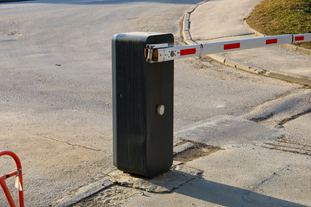 这张照片展示了一道安装在沥青路上的自动障碍物。障碍物的主体为灰色，配有一个白色按钮和一根带有红色条纹的白色栏杆。图片下载