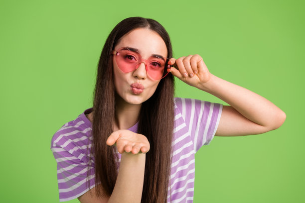 可爱的年轻女士的照片肖像，戴着心形太阳镜，送出空气之吻，穿着时尚的紫色条纹服装，背景为绿色。图片下载