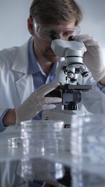 穿着实验室外套、白手套和防护眼镜的男性科学家在现代实验室中使用显微镜进行研究，肖像视图。低调照明照片。科学与医学图片下载