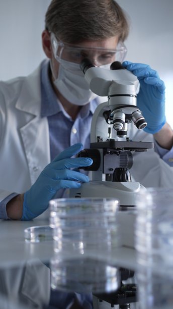 穿着蓝色手套、护目镜和医用口罩的男性科学家研究员正在现代实验室中使用显微镜，肖像视图。低调照明照片。科学与医学概念。图片下载