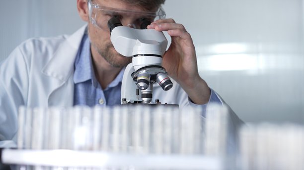 穿着实验室外套和防护眼镜的男性科学家在现代实验室中使用显微镜进行研究，肖像视图。低调照明照片。科学与医学概念图片下载