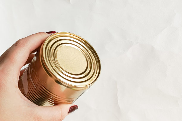 一只女性的手握着一个金色罐头，背景为白色，右侧留有文字位置。长保质期的罐装食品。罐头食品的宣传照片。图片下载