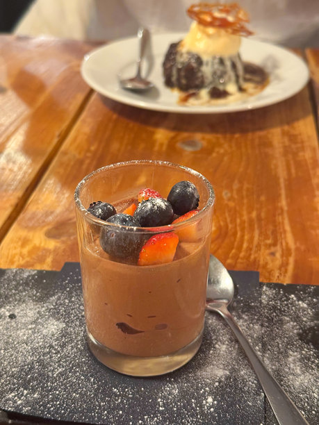 餐厅环境的图片，木纹桌子上摆放着一杯巧克力慕斯甜点，上面装饰着草莓和蓝莓，以及一盘粘性太妃糖布丁，配有香草冰淇淋和咸焦糖酱，重点在前景。图片下载
