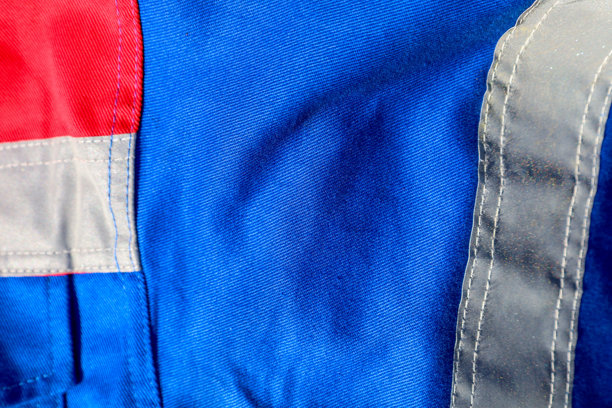 这张照片展示了一部分工作服的特写。照片中有蓝色面料，带有红色插片和灰色反光条。图片下载