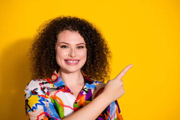 时尚的年轻女性在黄色背景下穿着色彩鲜艳的印花衣服，指向空白处的照片肖像。图片下载