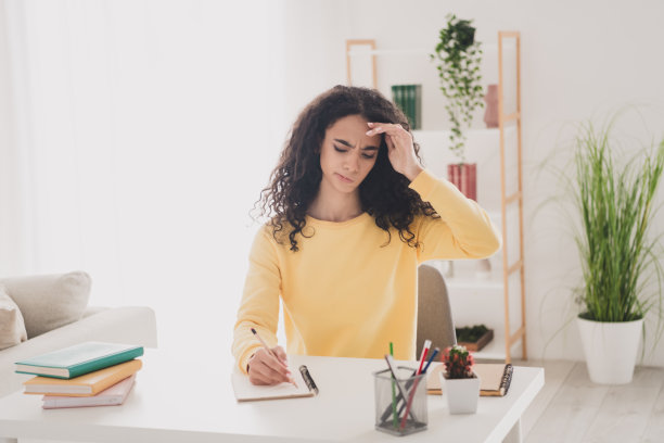 穿着黄色衬衫的美国女孩，年轻的波浪卷发，深思熟虑，坐在室内的工作场所做大学作业的照片肖像。图片下载