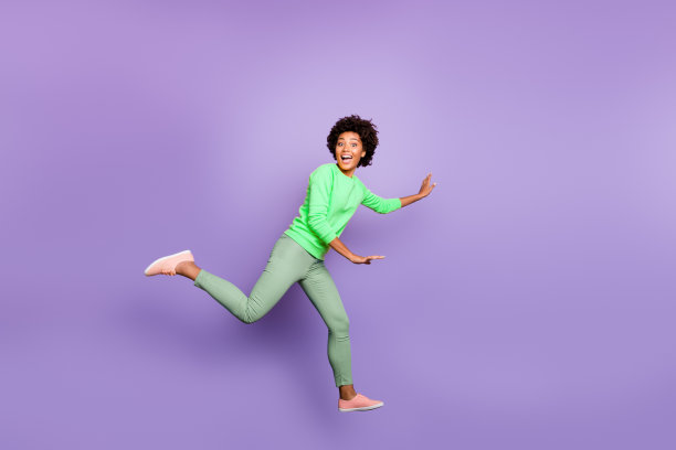全身侧面照片，展示一位开心兴奋的卷发混血女孩在春天与朋友们玩追逐游戏，穿着黄色毛衣和绿色裤子，背景为紫色。图片下载