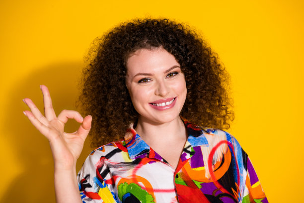 漂亮年轻女孩的照片肖像，做出“OK”手势，穿着时尚的彩色印花服装，背景为黄色。图片下载