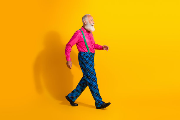 时尚老绅士穿着吊带裤走路的全身照片，背景为明亮的黄色。图片下载