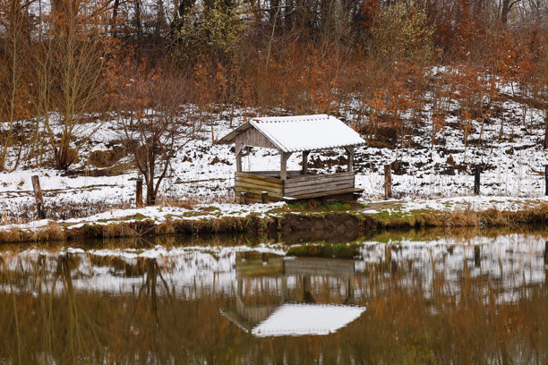 这张照片展示了一个小木凉亭，覆盖着雪，位于湖泊或池塘的岸边。凉亭的结构简单，拥有屋顶和开放的墙壁。图片下载