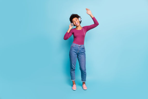全身照片，快乐积极的深肤色嬉皮女孩戴着耳机，唱着她最喜欢的立体声旋律，双手举起，穿着绿色毛衣和牛仔裤，背景为蓝色。图片下载