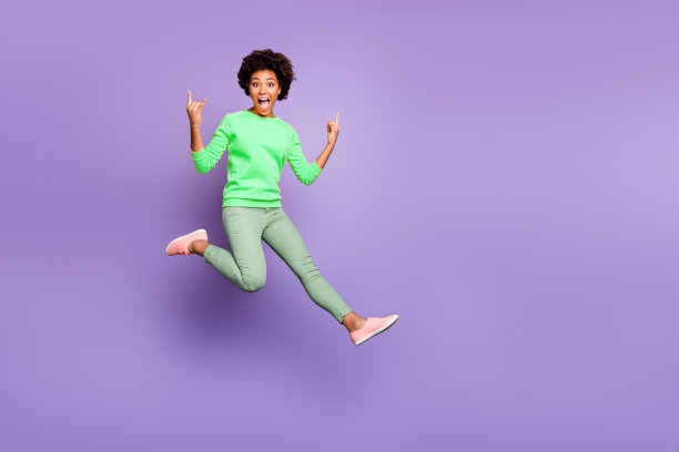 让我们摇滚！全身照片，快乐的疯狂混血女孩在秋春假期中展示角，做出真正的摇滚手势，穿着黄色毛衣和绿色裤子，背景为紫色。图片下载