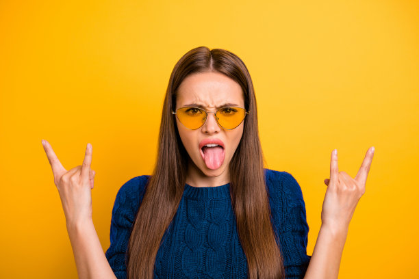 特写照片，快乐的女孩戴着眼镜，做出“牛角”手势，穿着酒红色毛衣，背景为黄色。图片下载