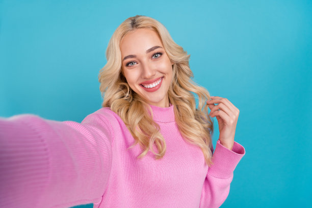 可爱的年轻卷发金发女孩穿着粉色毛衣在蓝色背景下自拍的照片图片下载