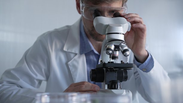 穿着实验室外套和防护眼镜的男性科学家在现代实验室中使用显微镜进行研究，肖像视图。低调照明照片。科学与医学概念图片下载