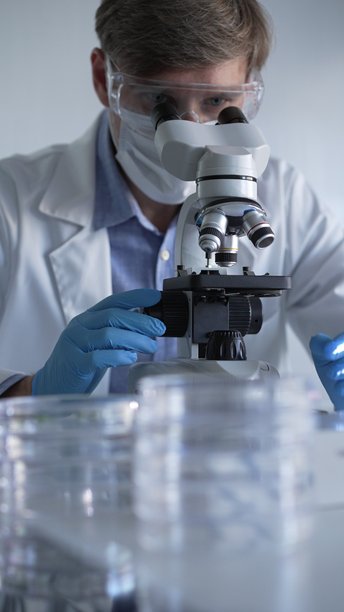 穿着蓝色手套、护目镜和医用口罩的男性科学家研究员正在现代实验室中使用显微镜，肖像视图。低调照明照片。科学与医学概念。图片下载