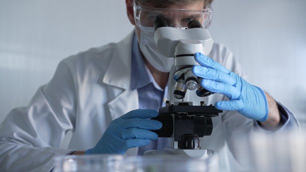 穿着蓝色手套、护目镜和医用口罩的男性科学家研究员正在现代实验室中使用显微镜，肖像视图。低调照明照片。科学与医学图片下载