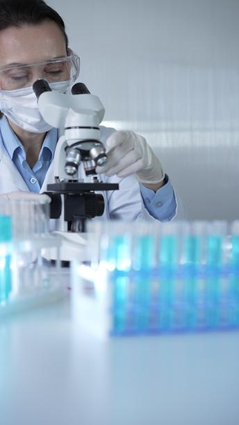 穿着实验室外套、戴着白色防护手套和眼镜的女科学家正在现代实验室中使用显微镜，肖像视图。低调照明照片。科学与医学概念。图片下载