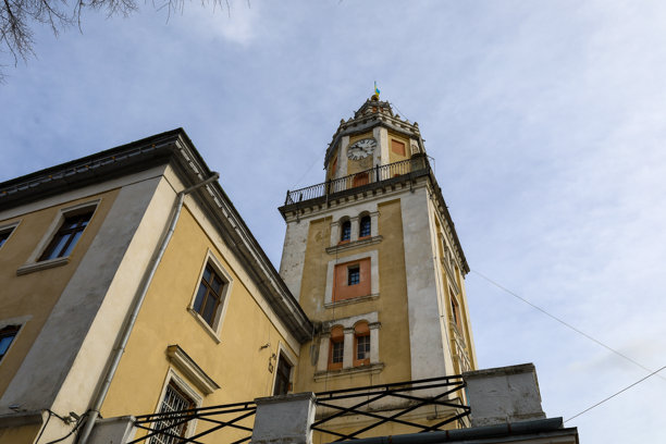 这张照片描绘了乌克兰桑比尔的历史市政厅建筑。从下方的视角可以看到一座高耸的钟楼和一个阳台，矗立在一栋有着黄色墙壁和棕色窗户的两层建筑上方。图片下载