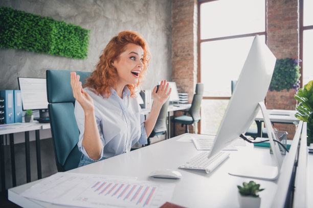 成功年轻女商人的个人资料照片，双臂举起，注视着电脑屏幕，现代阁楼风格的室内办公室。图片下载