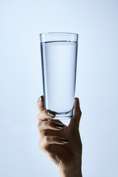 特写照片，修饰过的指甲的手握着装满水的透明玻璃，背景为柔和的蓝色。样机。图片下载