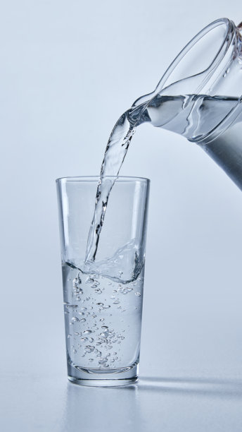透明水从水壶倒入透明玻璃杯的特写照片，展示气泡，背景为柔和的蓝色。模拟图。图片下载