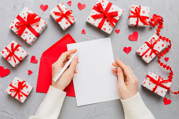 从上方拍摄的情人节装饰照片，女性手持信件和信封，旁边有小礼物盒和心形装饰，背景为单色柔和色调，留有空白空间。图片下载