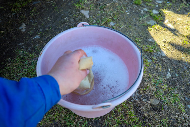 这张照片展示了一只手将海绵浸入一个粉色的肥皂水桶中。手属于一位穿着蓝色夹克的人，海绵呈奶油色。水桶中的肥皂水有白色泡沫和气泡。图片下载
