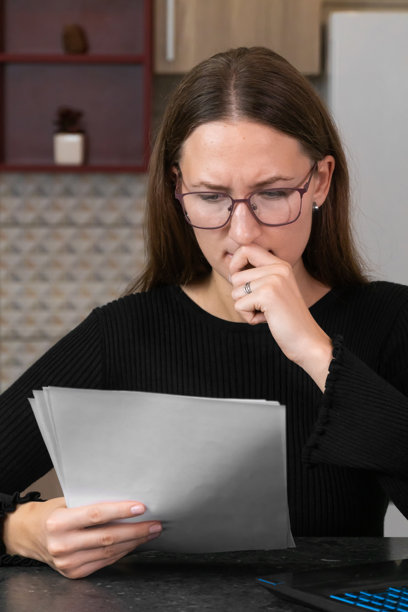 压力大的会计在繁忙的办公室环境中翻阅一堆会计文件，思考税务和财务问题，焦虑地用手触摸嘴唇。竖版照片图片下载