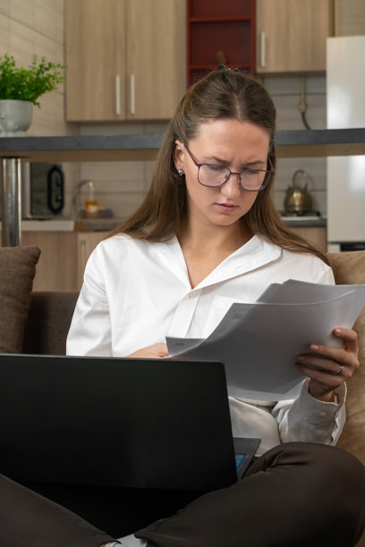 年轻女商人戴着眼镜，穿着白衬衫，坐在家中办公室的舒适沙发上，认真分析重要文件，同时在笔记本电脑上努力工作。竖版照片图片下载